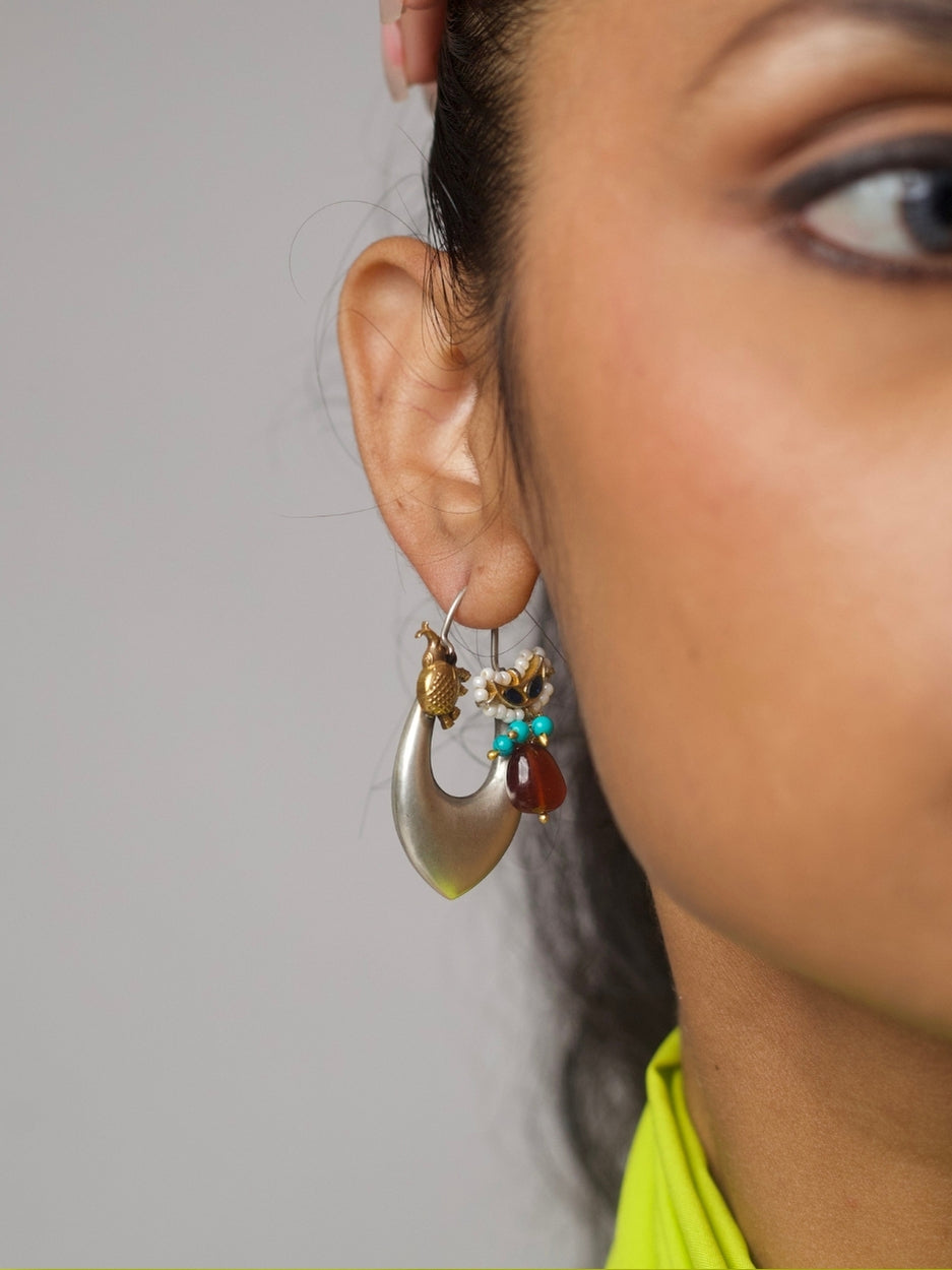 Elongated silver hoop earrings with gold bird motifs, pearls, turquoise beads, and deep red stone charms, styled on a lime green fabric.