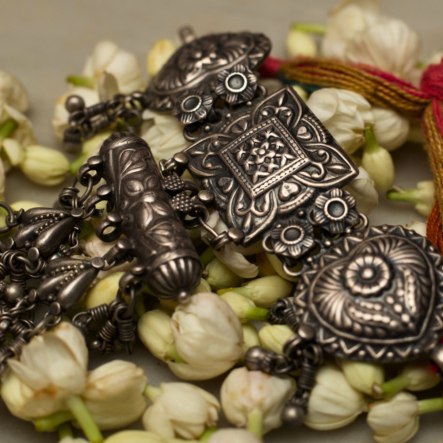 This unique traditional silver bracelet draws inspiration from the ancient hathphool. A delicate looking structure made strong and bold with pure 925 silver and the traditional method of oxidation. This bracelet features an embossed square centrepiece flaked by heart-shaped pieces attached to a red and green band with a flower-shaped closure. The most dreamy addition to this beauty is the cylinder and dangling beads attached to fall gracefully on your hands as you turn the pages of your favourite book.