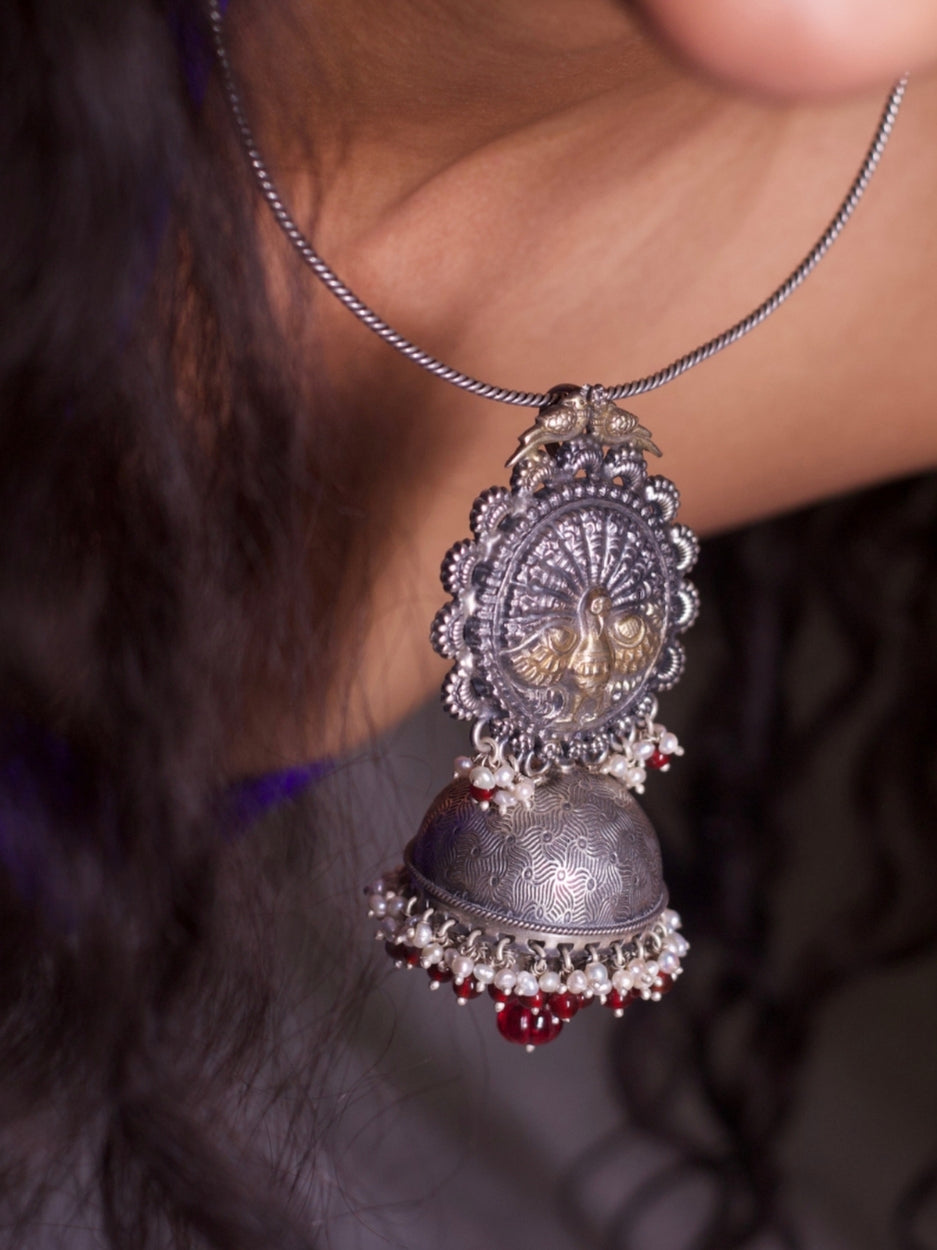Model wearing a handcrafted 92.5 silver torque necklace with a jhumka-style pendant, inspired by temple bells and tribal motifs. Features pearl and red bead detailing.