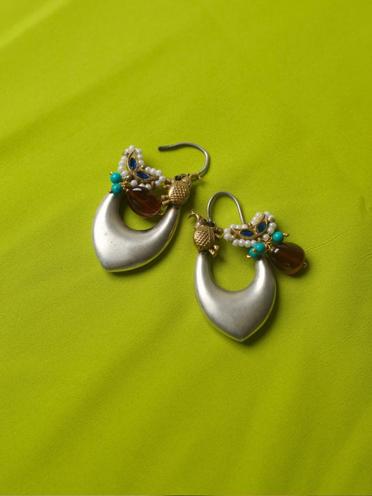 Elongated silver hoop earrings with gold elephant motifs, pearls, turquoise beads, and deep red stone charms, styled on a lime green fabric.