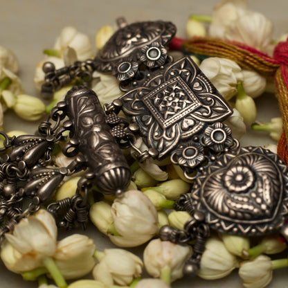 This unique traditional silver bracelet draws inspiration from the ancient hathphool. A delicate looking structure made strong and bold with pure 925 silver and the traditional method of oxidation. This bracelet features an embossed square centrepiece flaked by heart-shaped pieces attached to a red and green band with a flower-shaped closure. The most dreamy addition to this beauty is the cylinder and dangling beads attached to fall gracefully on your hands as you turn the pages of your favourite book.