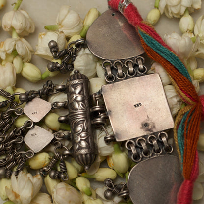 This unique traditional silver bracelet draws inspiration from the ancient hathphool. A delicate looking structure made strong and bold with pure 925 silver and the traditional method of oxidation. This bracelet features an embossed square centrepiece flaked by heart-shaped pieces attached to a red and green band with a flower-shaped closure. The most dreamy addition to this beauty is the cylinder and dangling beads attached to fall gracefully on your hands as you turn the pages of your favourite book.