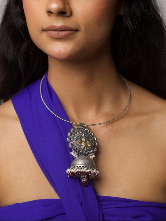 Model wearing a handcrafted 92.5 silver torque necklace with a jhumka-style pendant, inspired by temple bells and tribal motifs. Features pearl and red bead detailing.