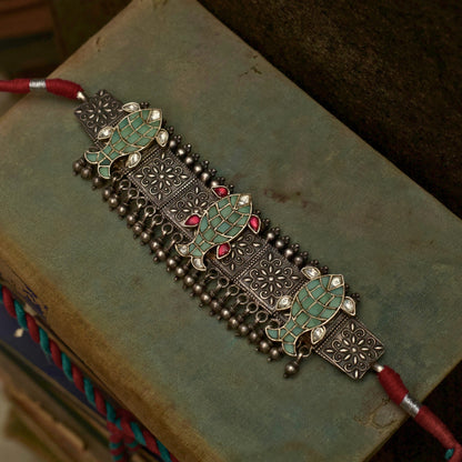This oxidised 92.5 silver choker features three meticulously designed fish cutouts in lush pastel green, with scarlet and white gemstones on an embossed base flanked by ghunghroo beads. It comes with a shiny pink, adjustable dhaaga that perfectly complements the colours and adds a vintage charm.