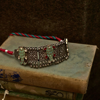 This oxidised 92.5 silver choker features three meticulously designed fish cutouts in lush pastel green, with scarlet and white gemstones on an embossed base flanked by ghunghroo beads. It comes with a shiny pink, adjustable dhaaga that perfectly complements the colours and adds a vintage charm.