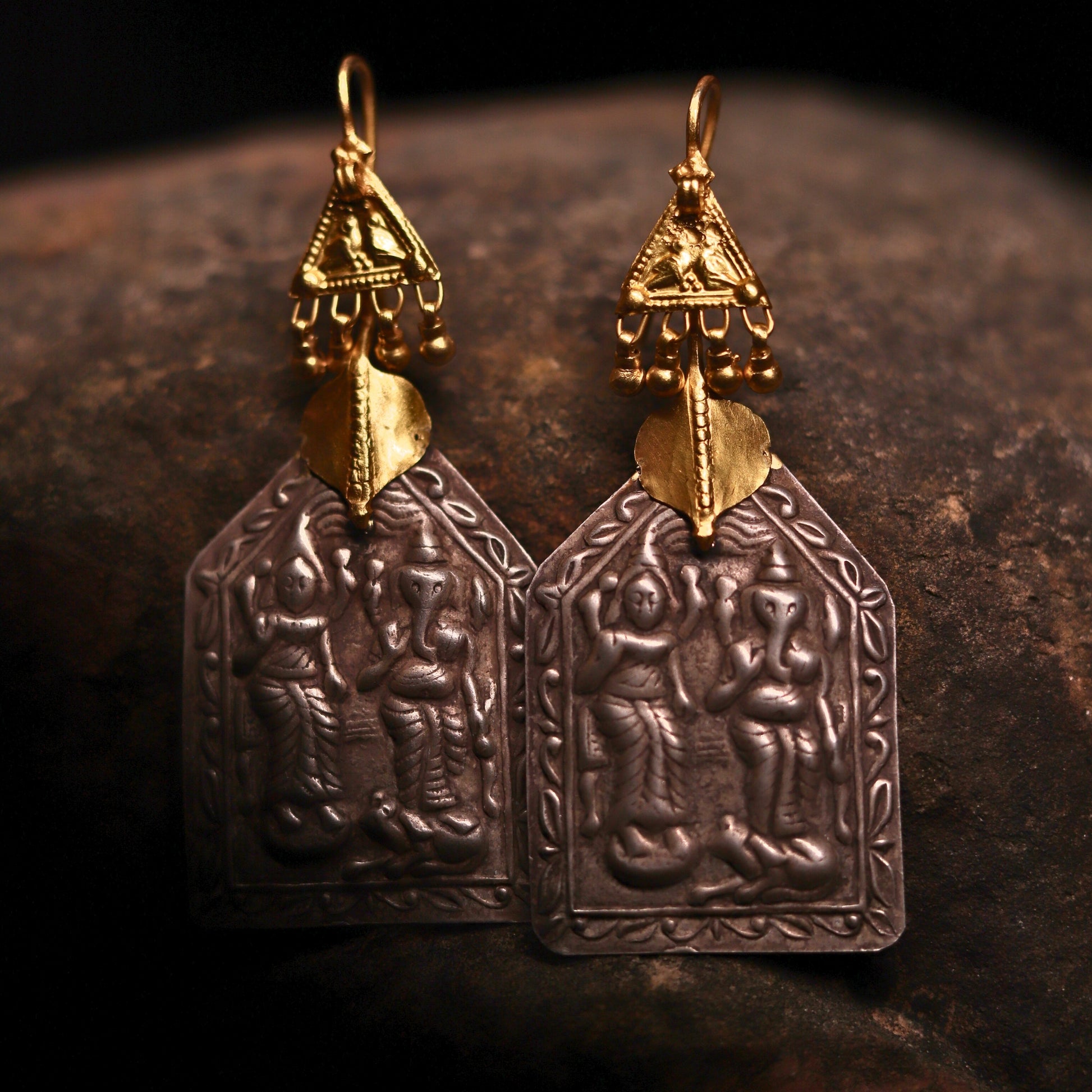 Silver earrings featuring Goddess Laxmi and Lord Ganesha embossed on antique silver pentagon plates, paired with gold-polished triangles and dangling beads resembling a mythical chariot