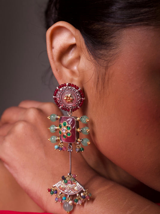 Layered silver statement earrings with red circular stud, oxblood enamel rectangle with green kundan floral motifs, and fan-shaped jharokha drop adorned with multicolor gemstone beads.